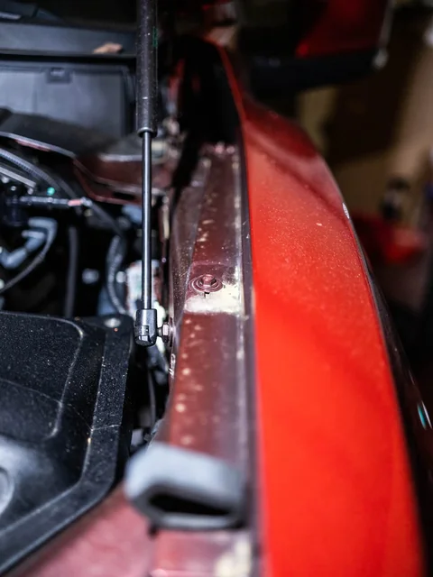 A hood strut beside the edge of a red fender, hood up