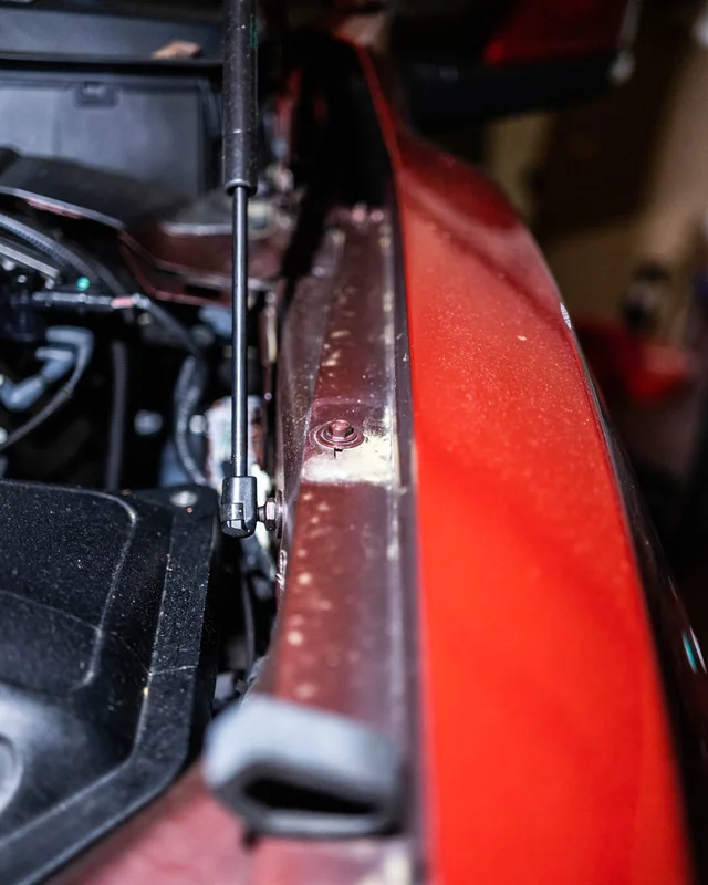 A hood strut beside the edge of a red fender, hood up